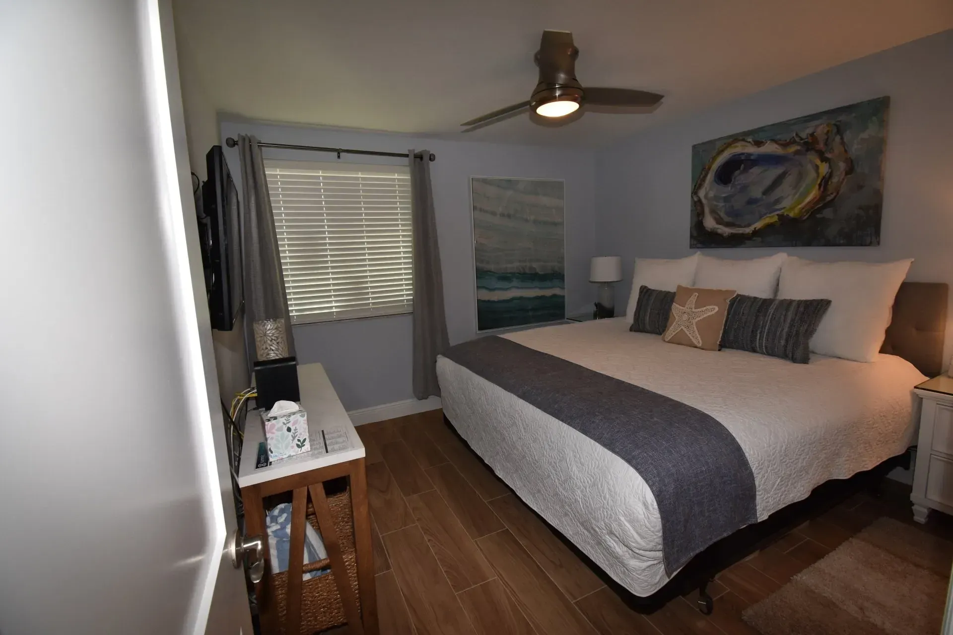 Bedroom with a queen bed, art, ceiling fan, and window. Pale blue walls, wooden floors, and white bedside table.