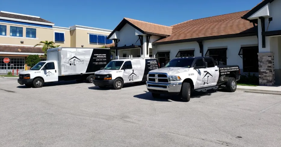 Three white work vehicles parked in a lot, with a company logo on the sides, in front of a building with brown roof.