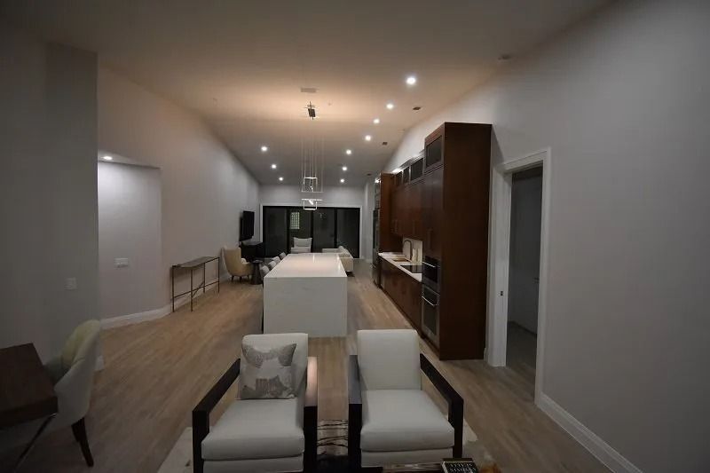 Modern, spacious kitchen/living area with white island, wooden cabinetry, and light wood flooring.