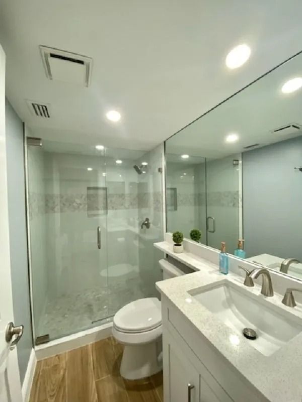 Bathroom with glass shower, white vanity, toilet, and large mirror; light blue walls, wood-look floor.