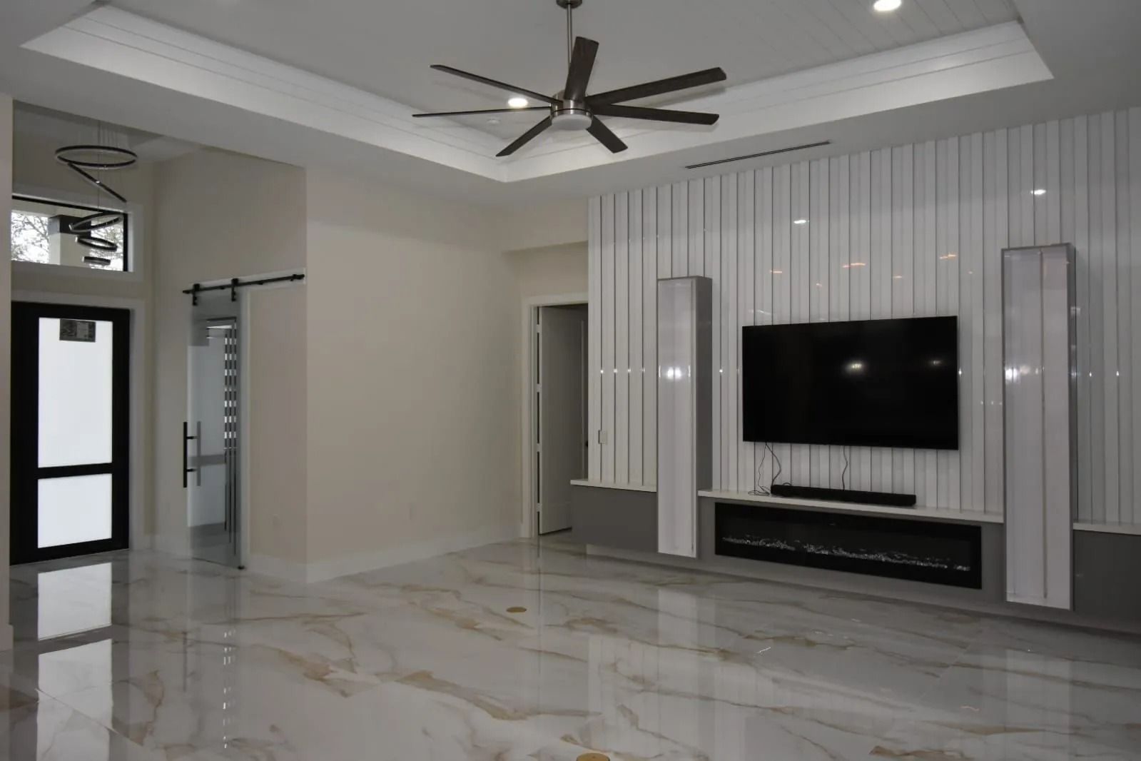 Spacious, modern living room with white walls, marble floors, large TV, fireplace, and a ceiling fan.