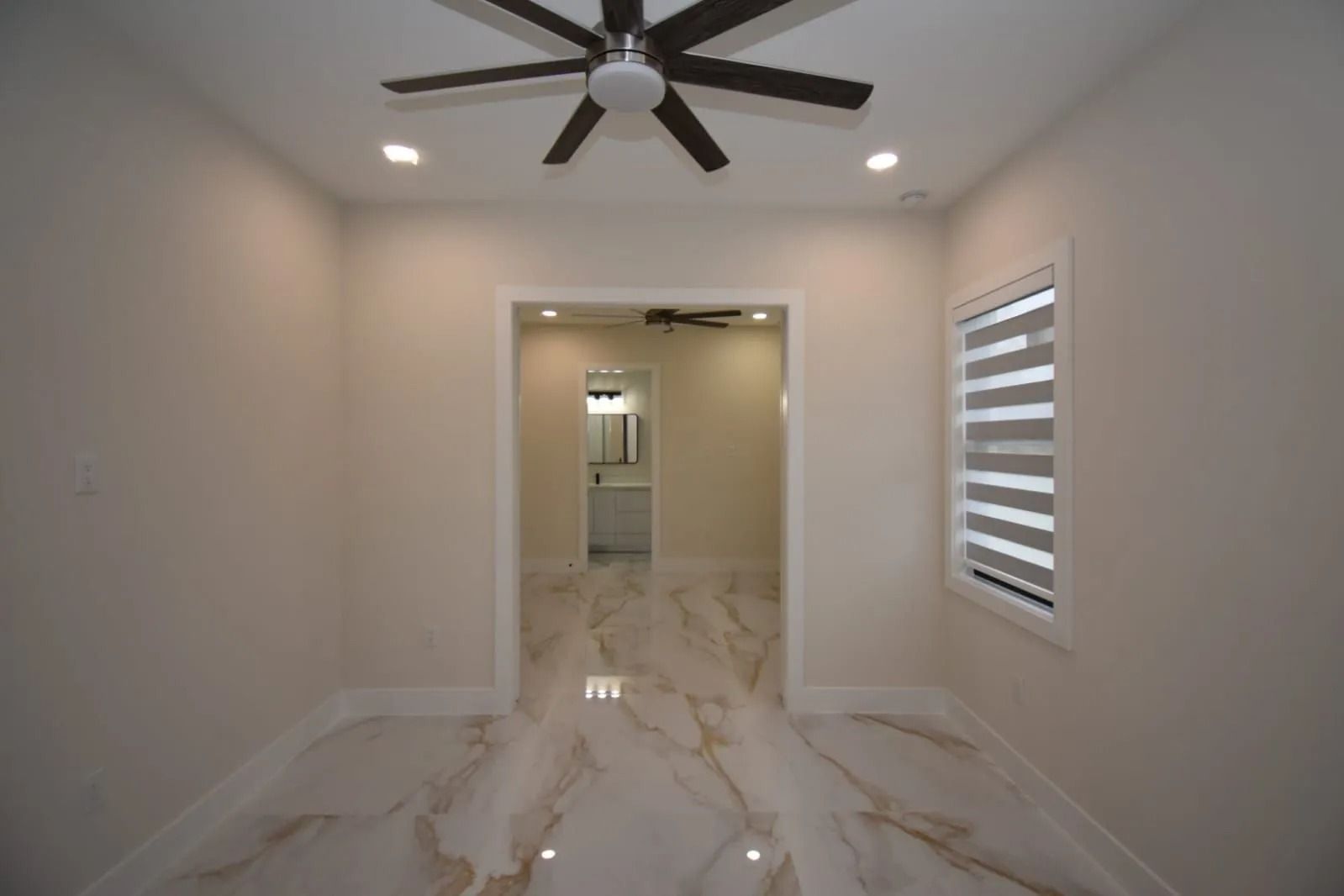Empty room with marble floors, leading to a hallway, with a ceiling fan and a window with blinds.