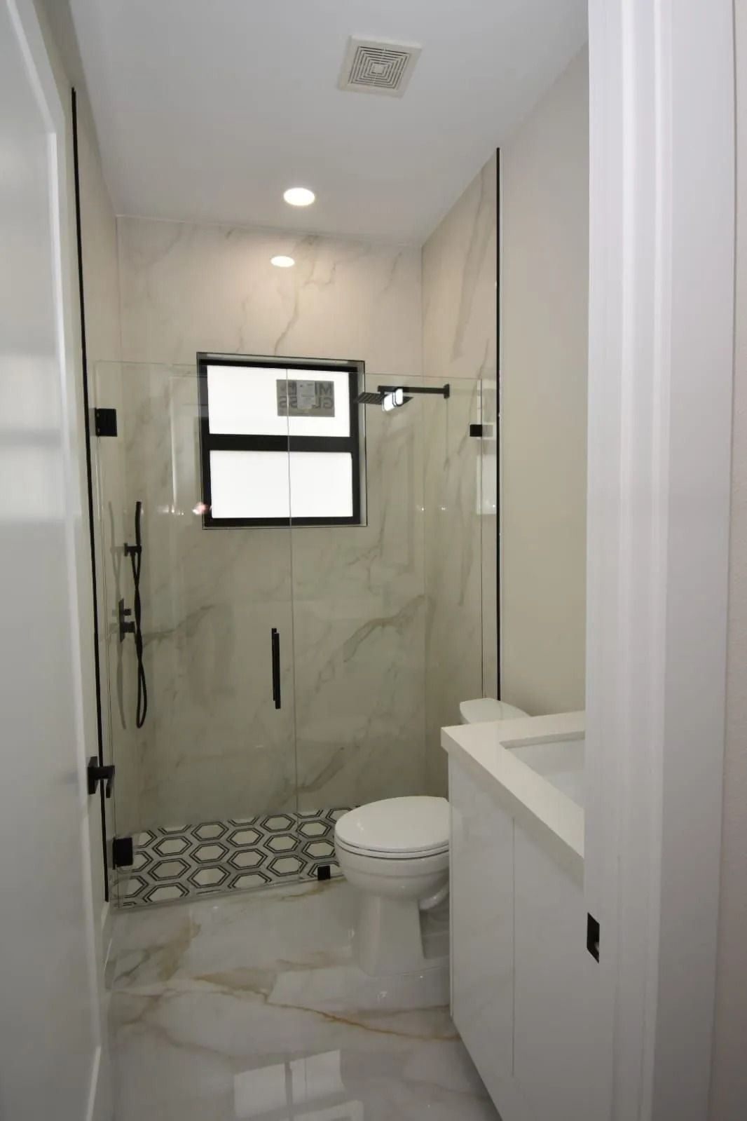 Modern bathroom with marble walls, glass shower, white vanity, toilet, and black accents.