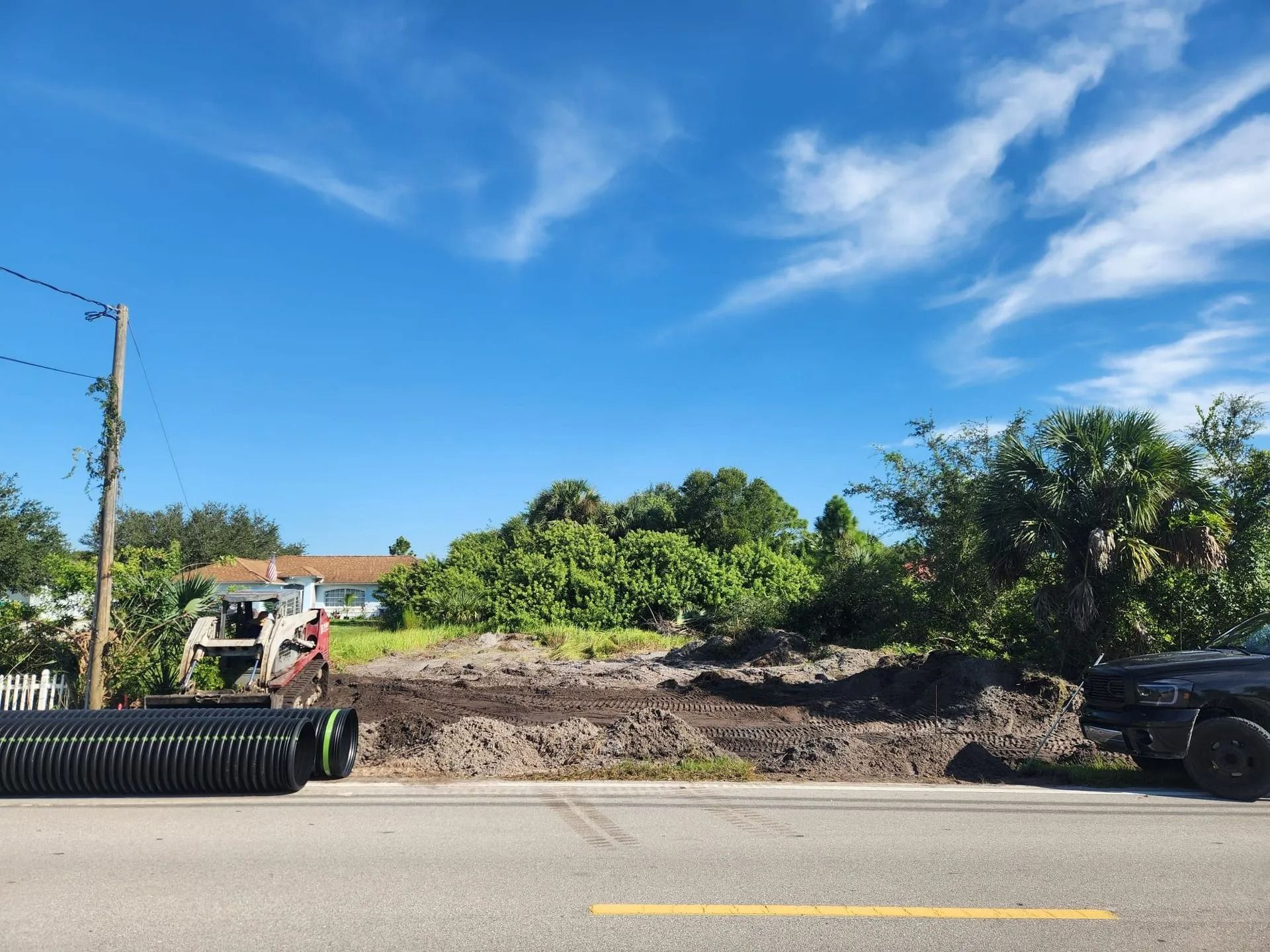 Construction site with exposed earth, shrubs, and sky; black corrugated pipes sit on the left.