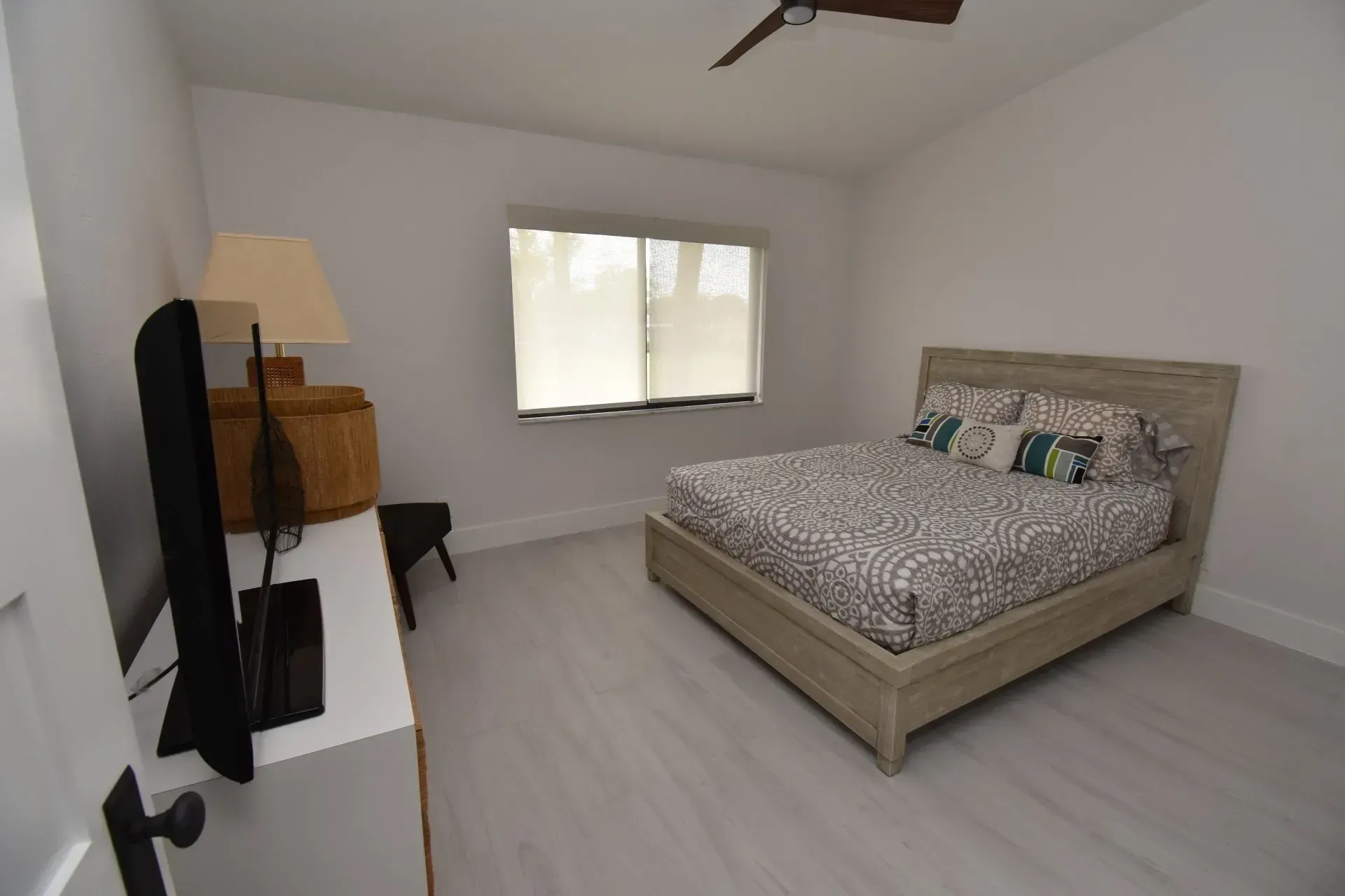 Bedroom with bed, lamp, TV, and window. Light wood floor and neutral walls.