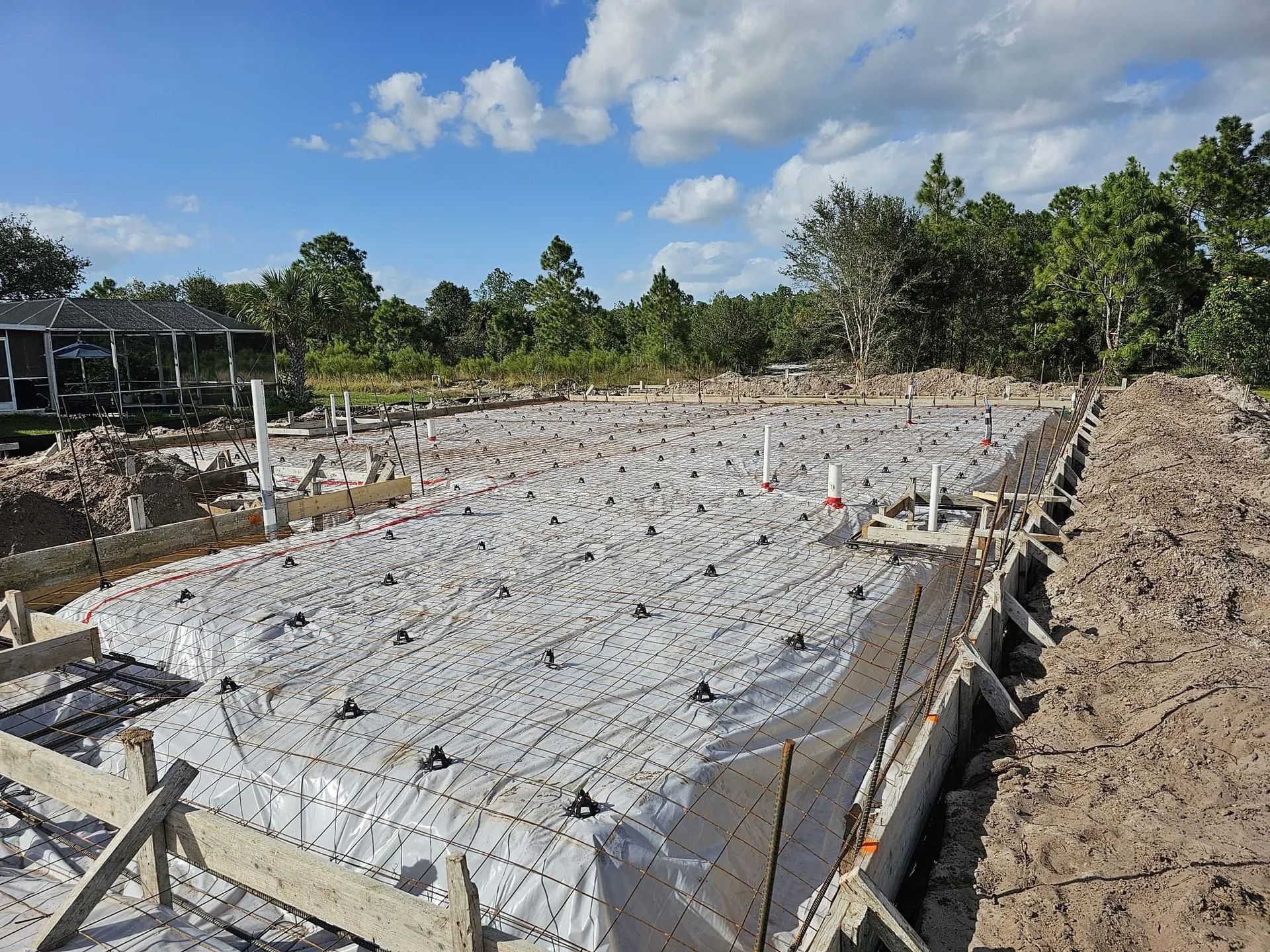 Construction site with concrete foundation, white plastic cover, pipes, wood forms, and surrounding trees under a cloudy sky.