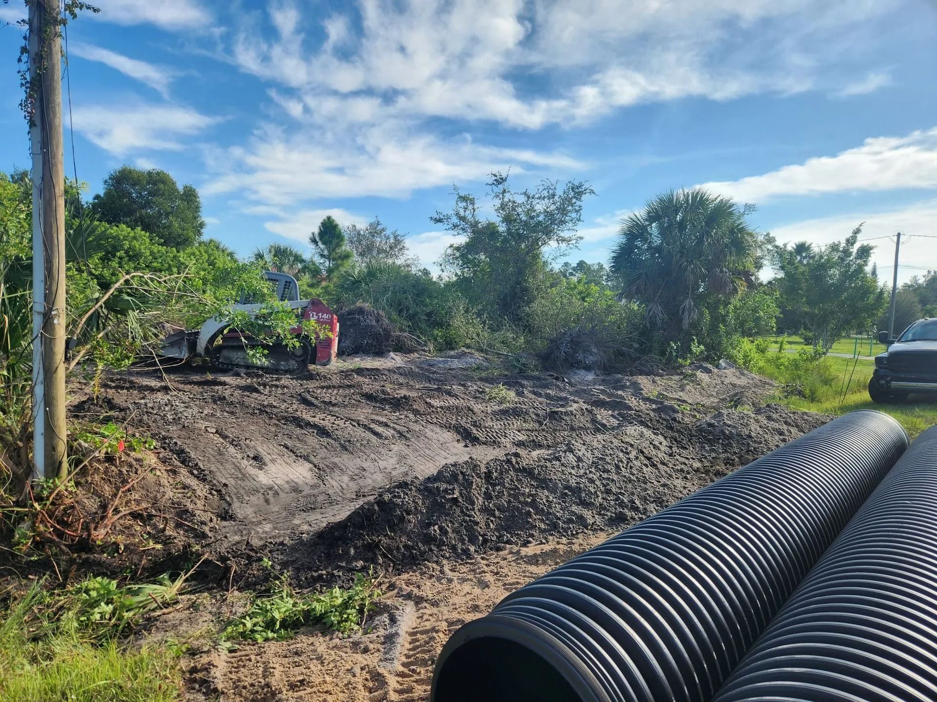 A construction site with dirt piles, black drainage pipes, and greenery under a cloudy sky.