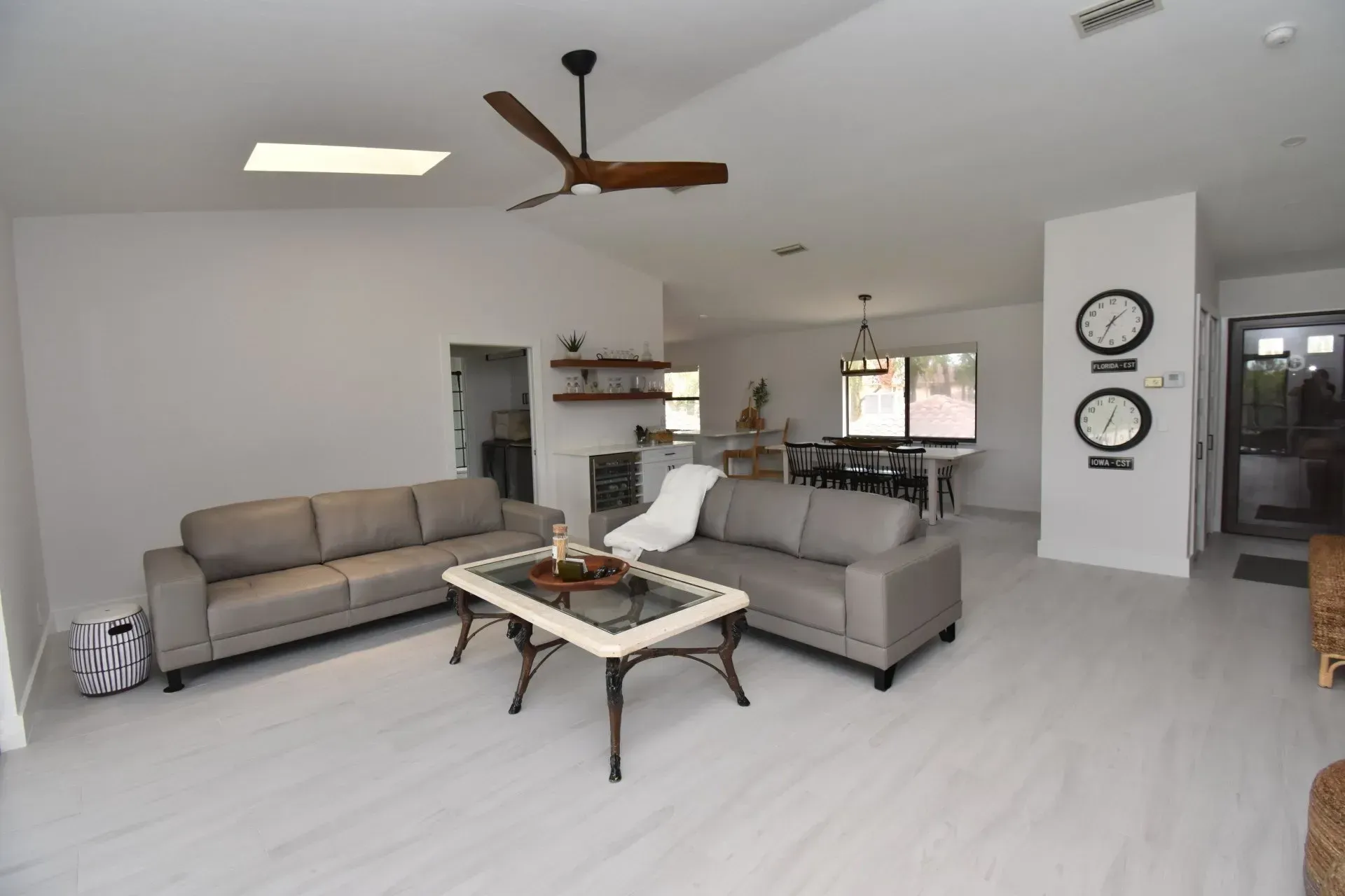Living room with gray sofas, a coffee table, and a dining table, featuring white walls, a ceiling fan, and clocks.
