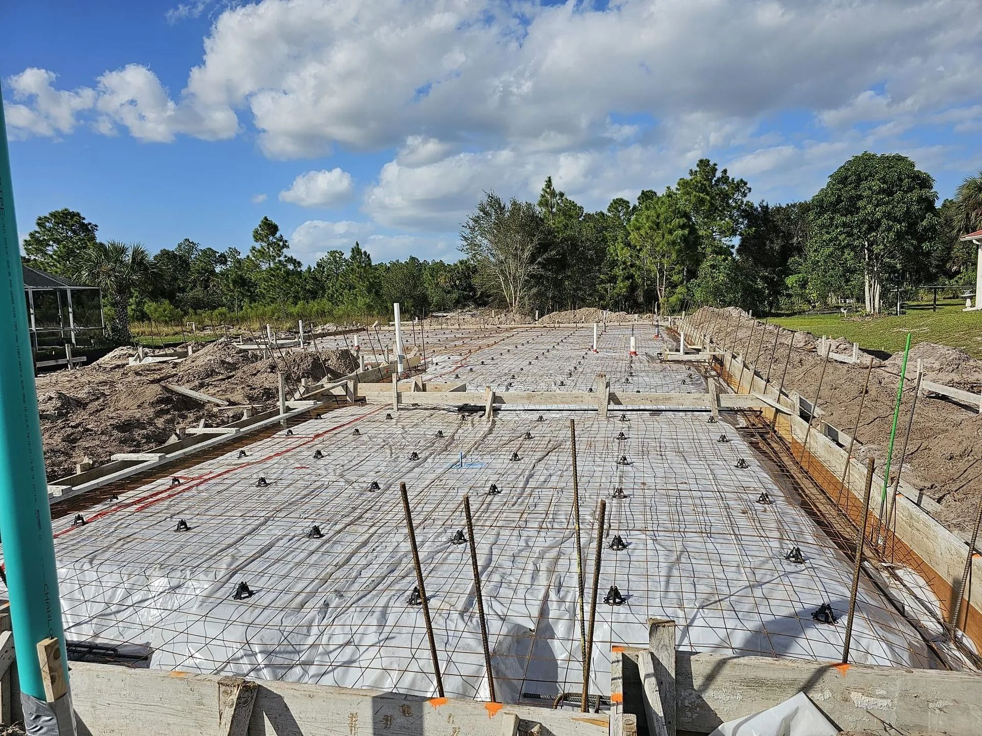 Construction site with foundation laid; rebar, pipes, and white vapor barrier visible. Cloudy sky.