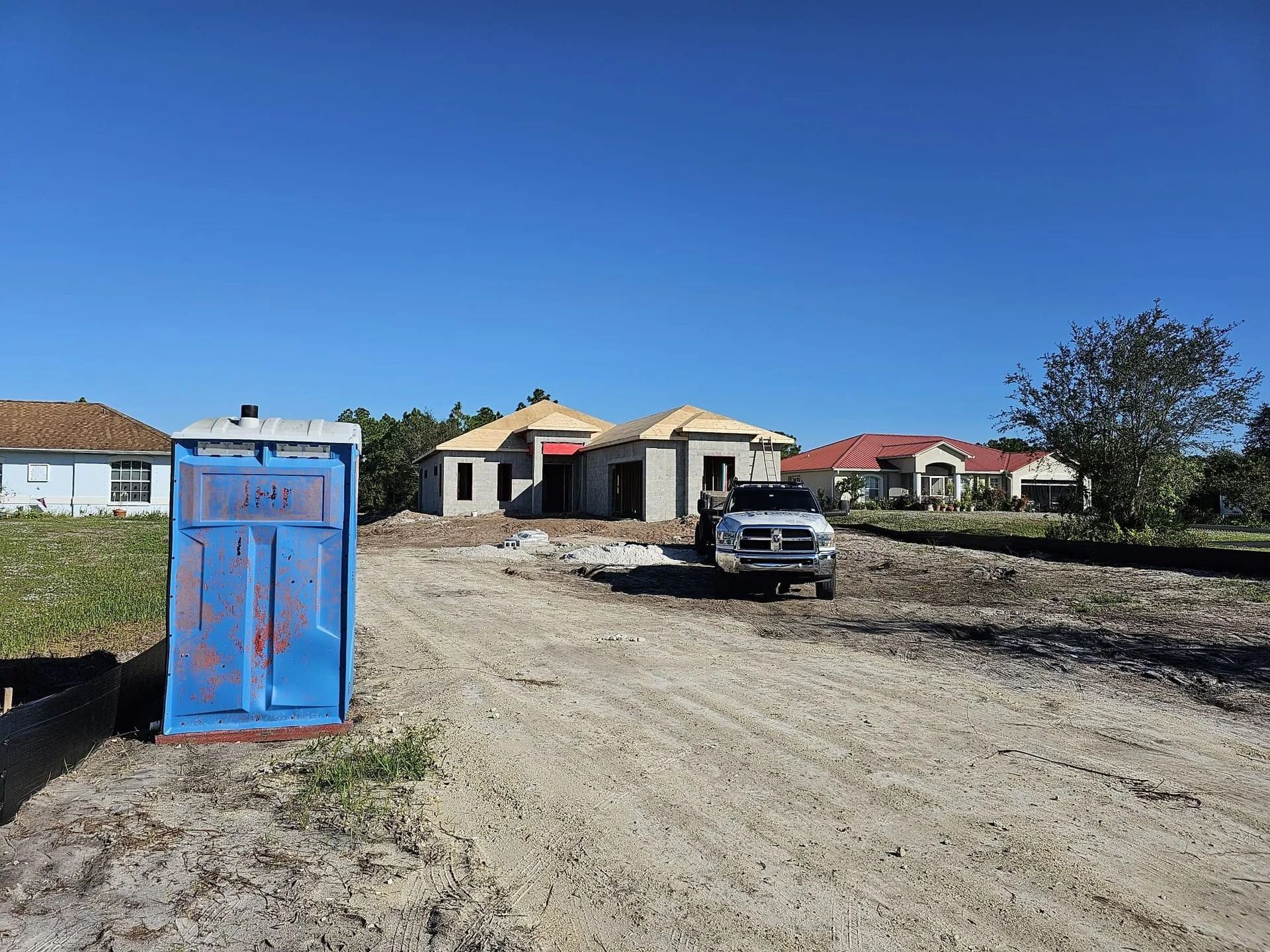 Blue portable toilet sits on a construction site with a truck and house under construction on a sunny day.