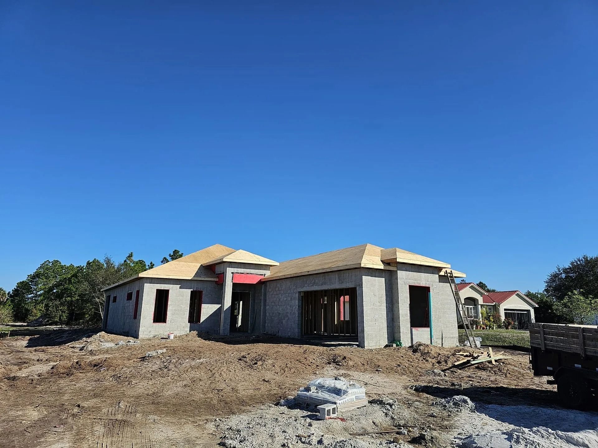 House under construction, gray concrete walls, wood roof frame, blue sky, dirt lot.