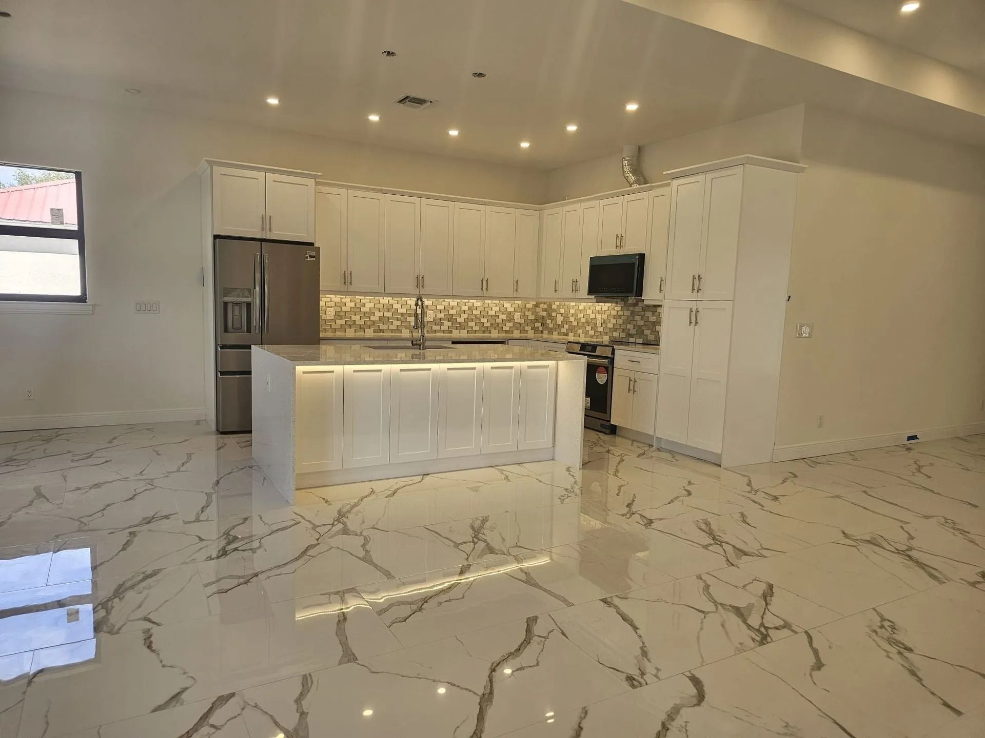 Modern white kitchen with stainless steel appliances, large island, and marble-look flooring.