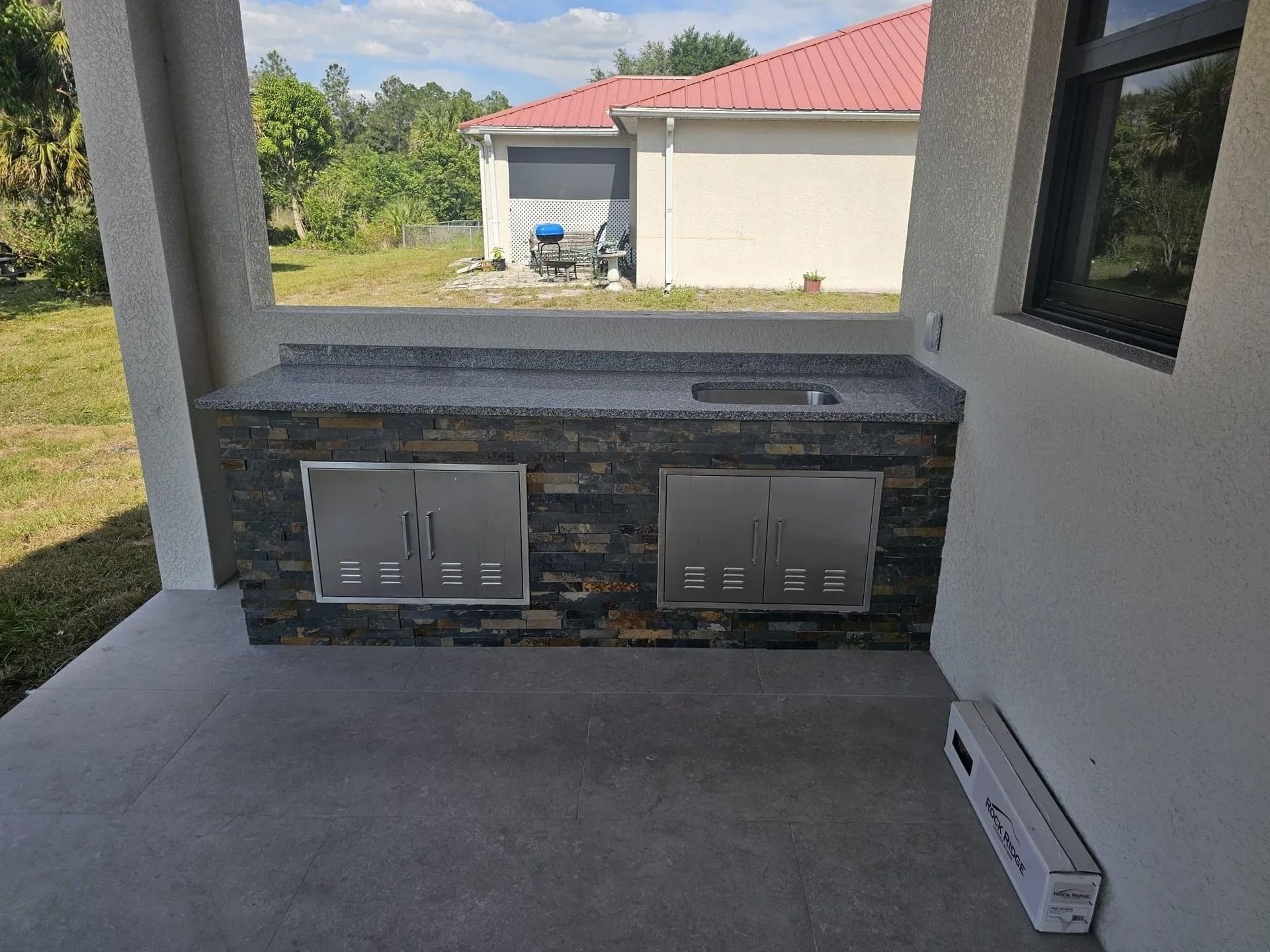 Outdoor kitchen with stone facade, gray countertop, stainless steel doors, and sink.