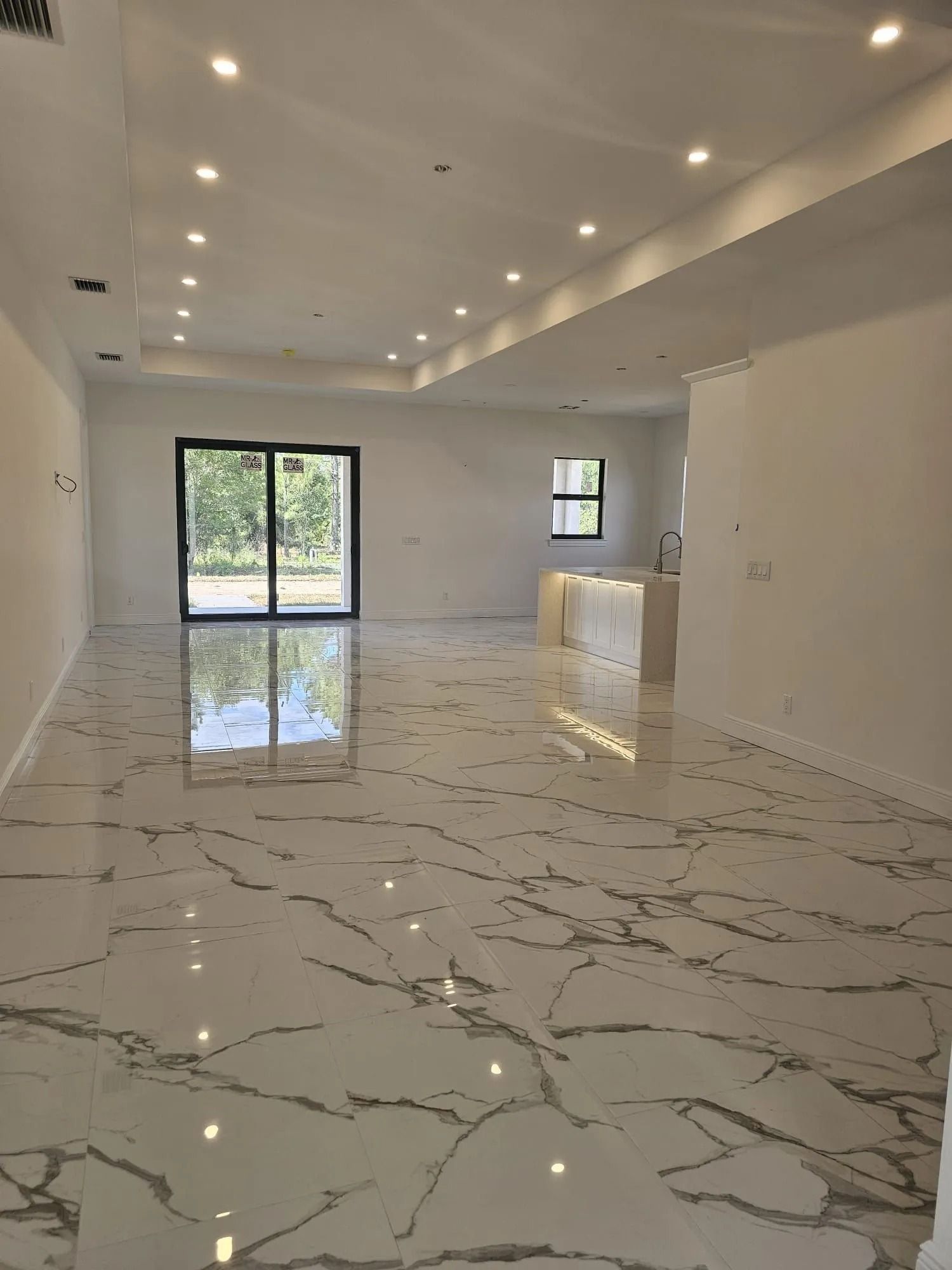 Empty living room with white marble-look floors, recessed lighting, and sliding glass doors to a backyard.