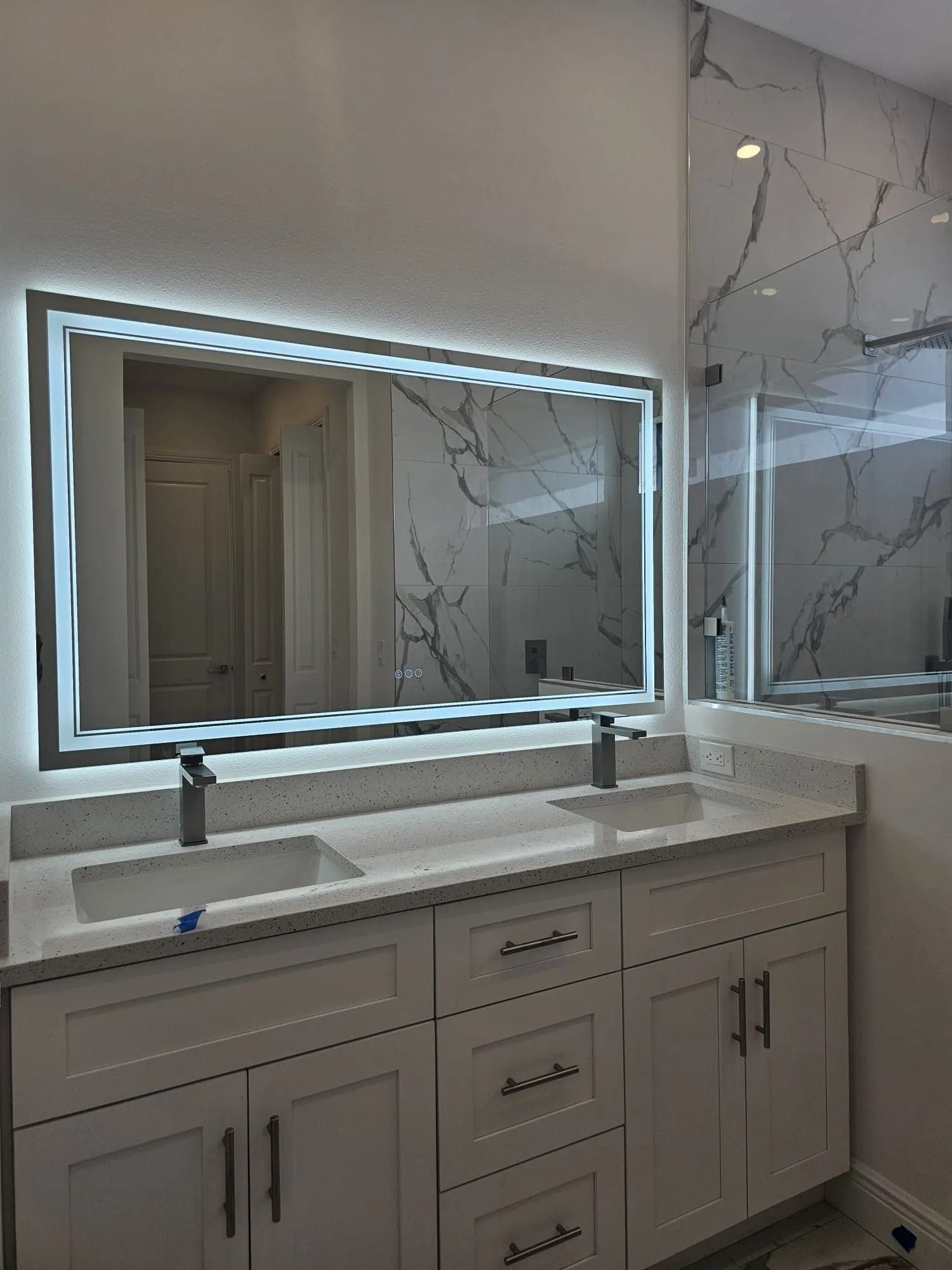 Bathroom with white cabinets, marble-look wall, large illuminated mirror, and double sink.