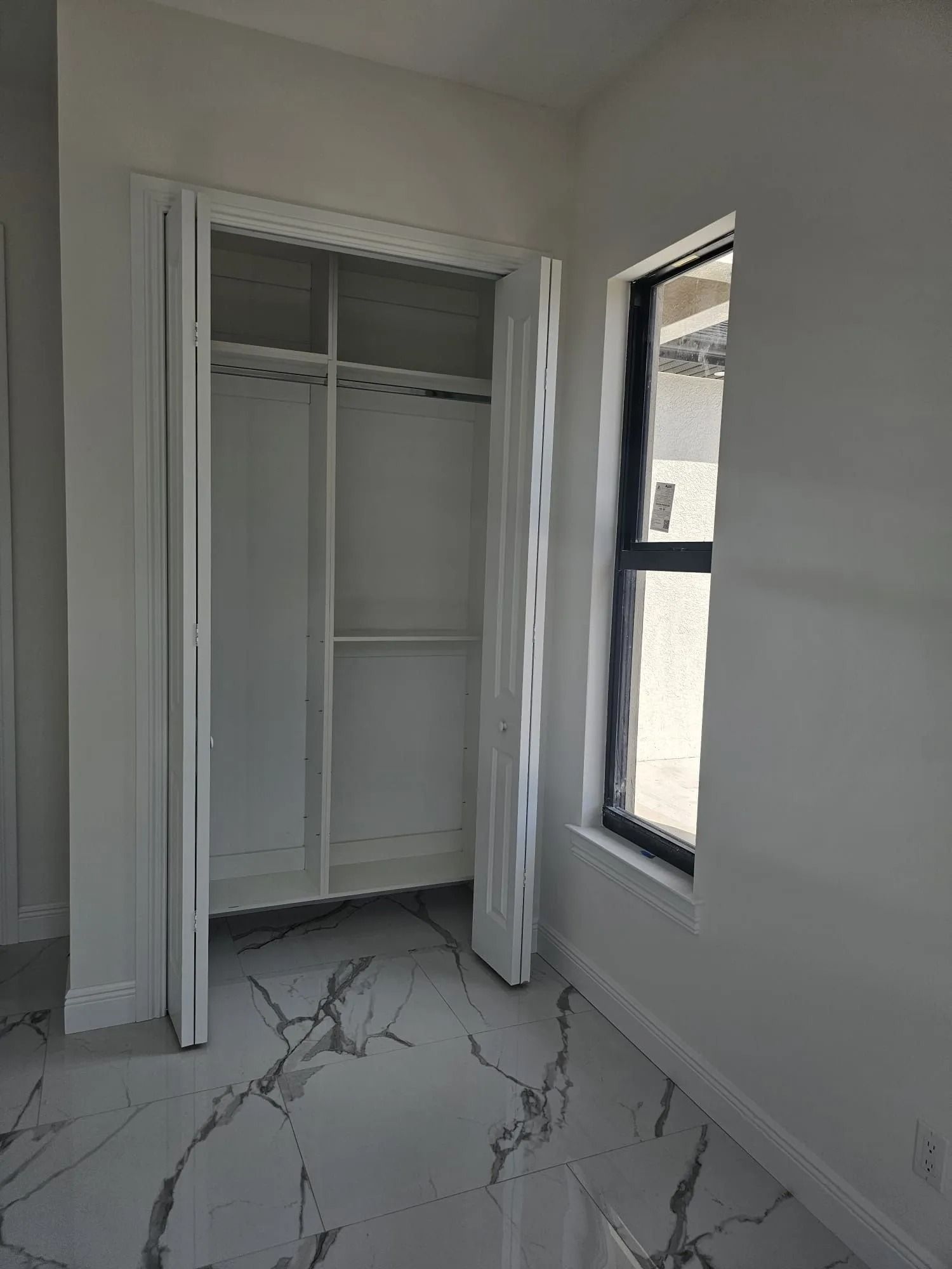 White closet with bi-fold doors, white walls, and a window with a black frame. Marble-look floor.