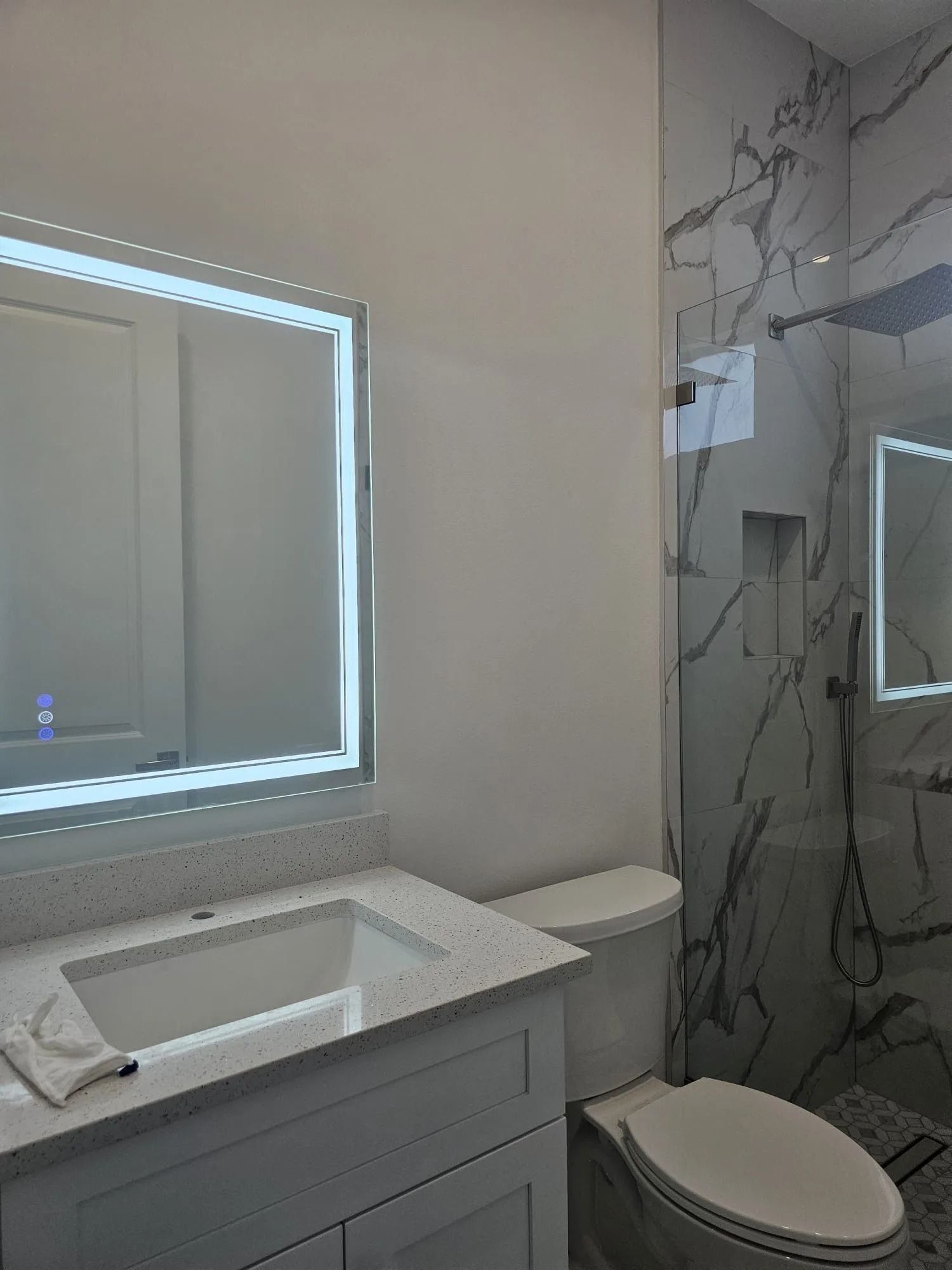 Bathroom with a backlit mirror, marble shower, white vanity, and toilet.