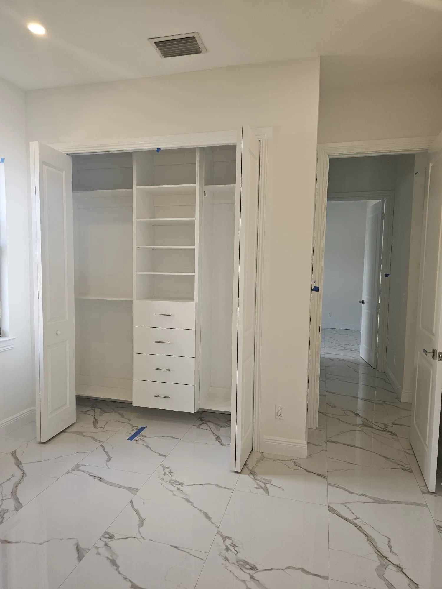 White walk-in closet with built-in shelves and drawers, next to an open doorway with marble flooring.