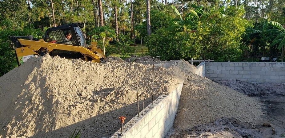 A yellow skid steer moving sand at a construction site near a wall and trees.
