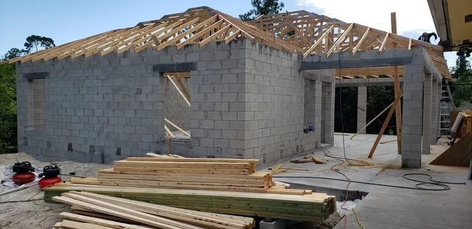 House under construction with block walls and wooden roof supports. Lumber is piled in foreground.