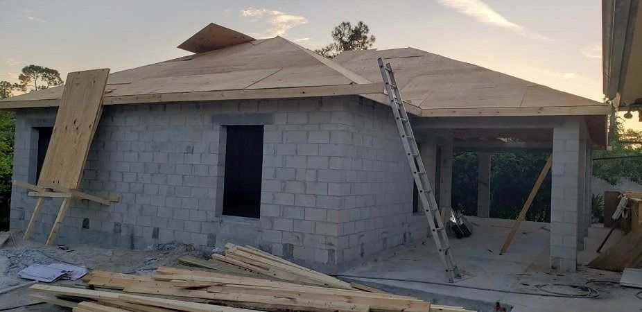 House under construction, concrete block walls, plywood roof, ladder, wood materials scattered.