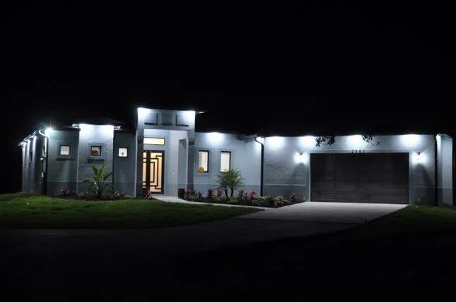 A modern home at night, illuminated by outdoor lights; dark driveway and black sky.