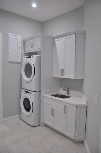 White laundry appliances stacked, next to a corner sink with white cabinets and a wall-mounted cabinet.