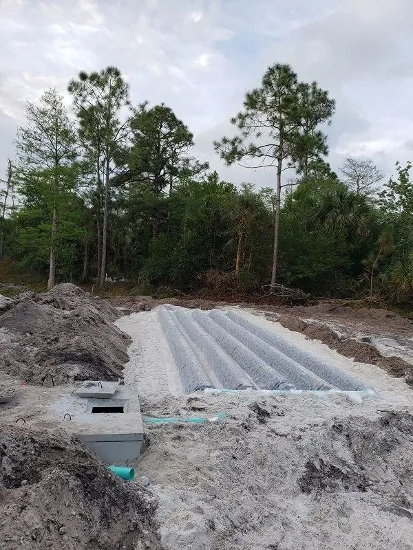 Septic system under construction in a sandy clearing, with grey concrete tank and distribution field pipes.