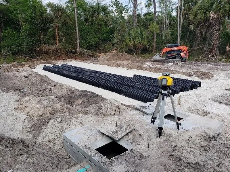 Septic system installation: black drainage pipes on a sand base with a laser level and mini excavator.