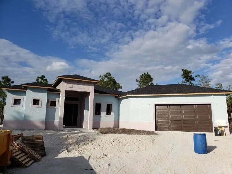 House under construction, pale blue stucco exterior, brown garage door, blue sky.