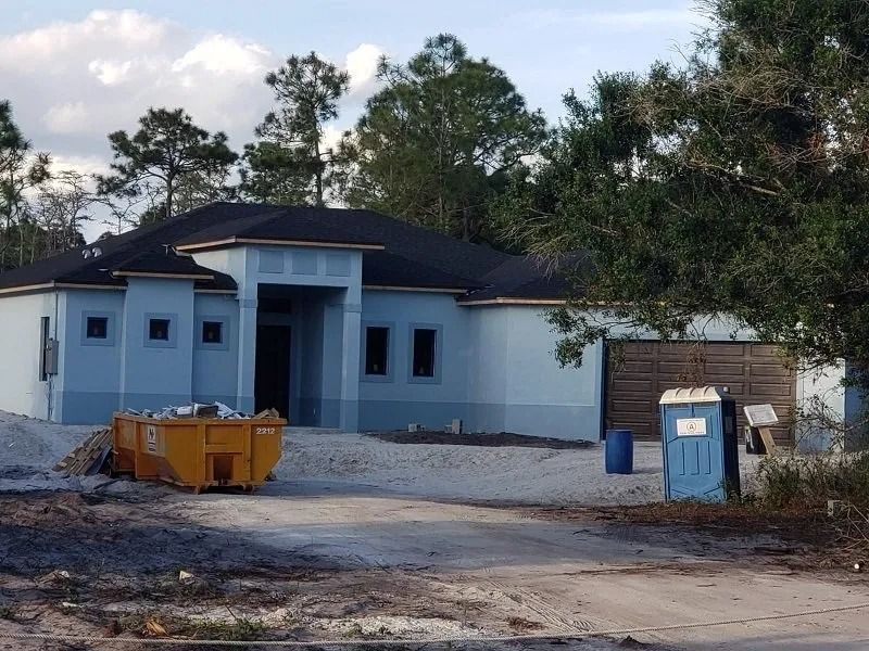 A light blue house under construction with a dark roof, construction debris, and a portable toilet in the yard.