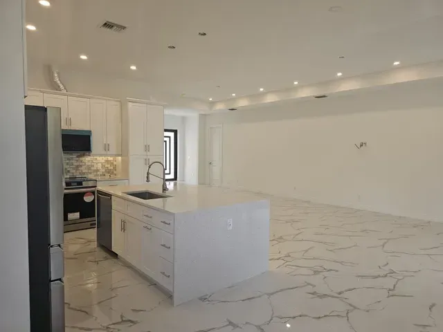 Modern kitchen with white cabinets, island, and marbled floor. Empty living space with recessed lighting.