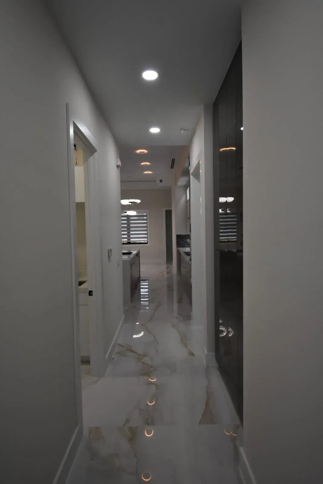 Narrow hallway with marble floors and recessed lighting, leading to a kitchen with blinds.