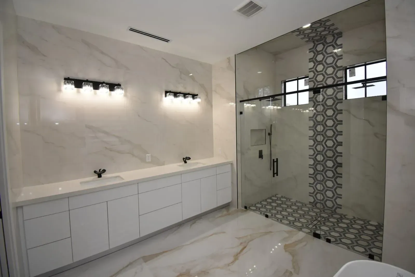 Modern bathroom with white marble tile, double vanity, glass shower, and black light fixtures.