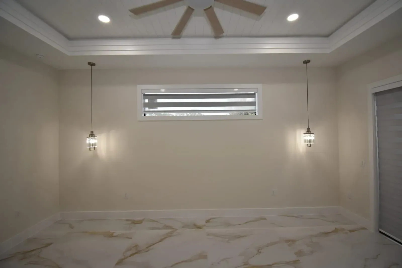 Empty, beige-walled room with marble floors, a rectangular window, and two hanging lamps. A ceiling fan is visible.