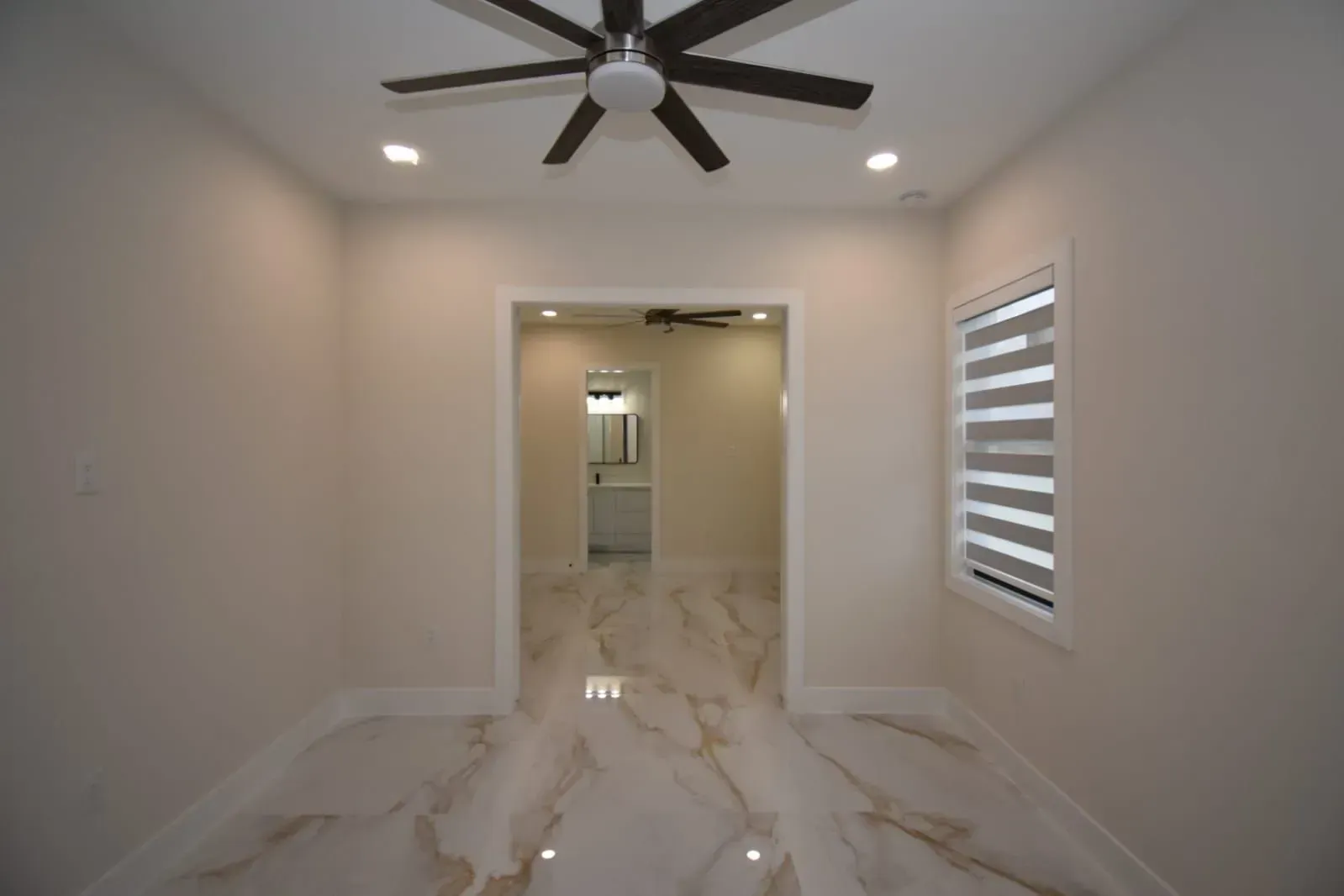 Empty room with marble floors and recessed lighting, leading to a doorway, and a window with blinds.