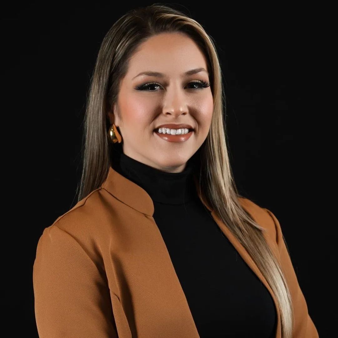 Woman in a black turtleneck and tan blazer smiles against a black backdrop.