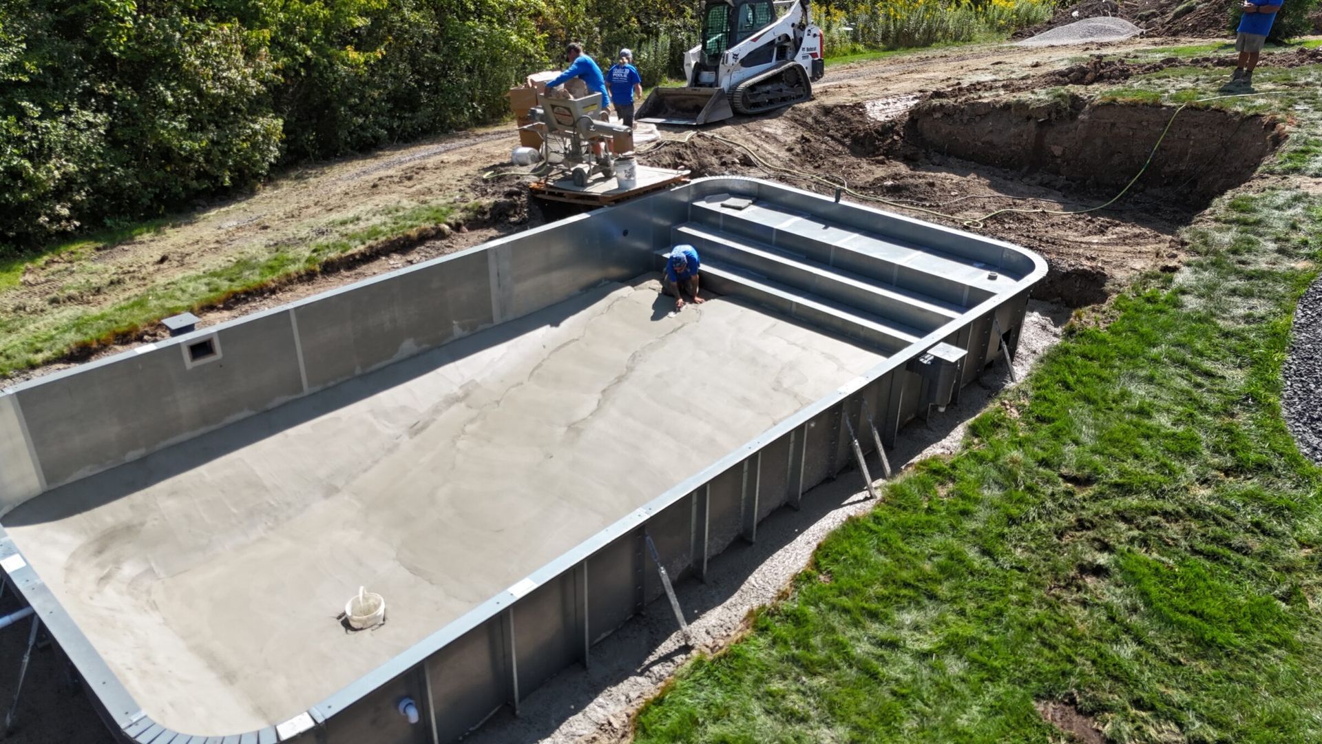 A partially constructed concrete pool with visible steel framing, as a worker smooths out the concrete base. Construction equipment and materials are placed nearby on a grassy lot.