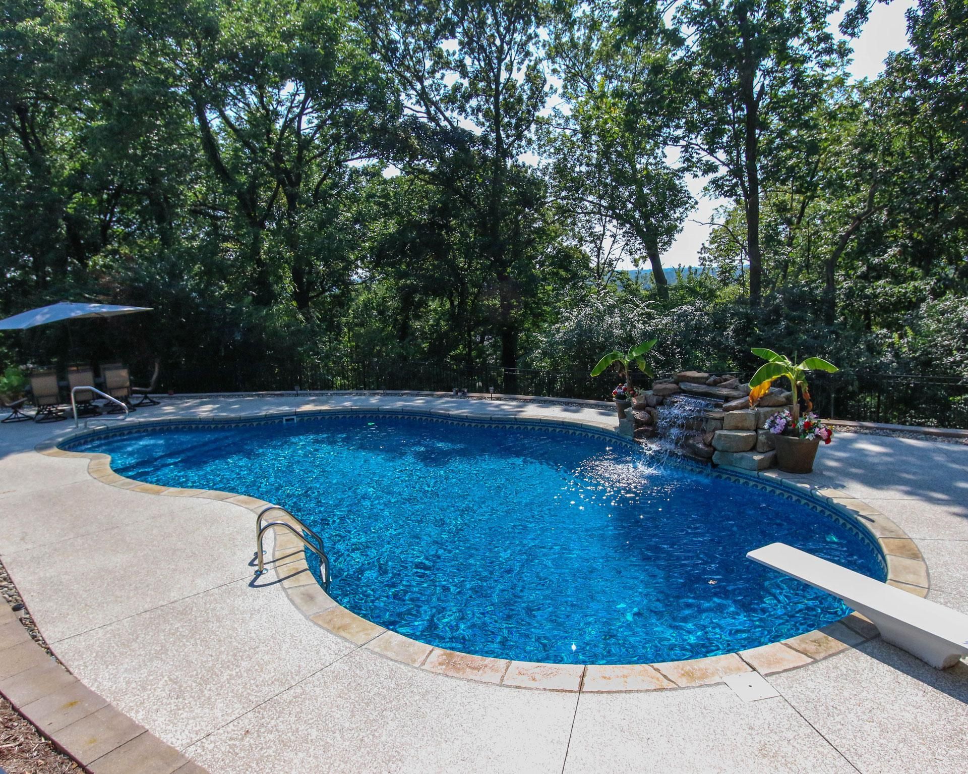 A curved, freeform backyard pool with a white diving board and a stone waterfall feature. The pool is surrounded by trees and shaded seating areas, giving a natural, private retreat feel.