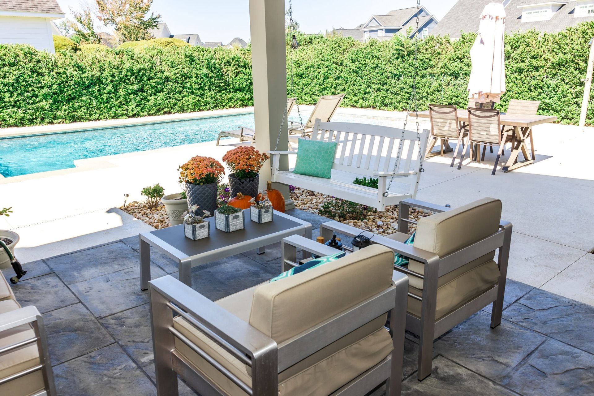 Covered patio seating area with cushioned chairs, a white porch swing, and fall-themed potted plants overlooks a rectangular backyard pool and lounge area.