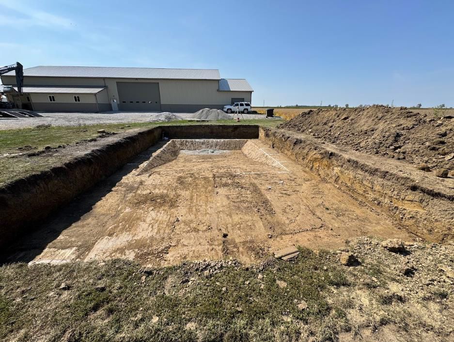 A rectangular excavation in a field, with a large building in the background. Sunny day.