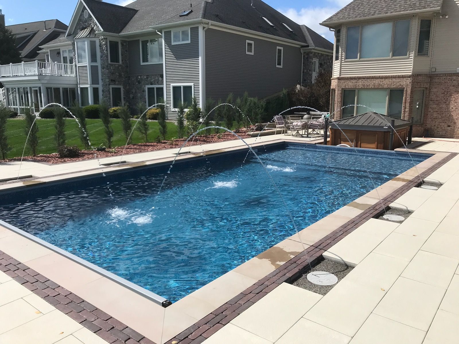 Rectangular pool with water fountains, surrounded by tan patio, near houses with landscaping.