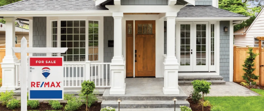 A gray suburban house with a wooden front door, white porch pillars, and a RE/MAX For Sale sign in the front yard.