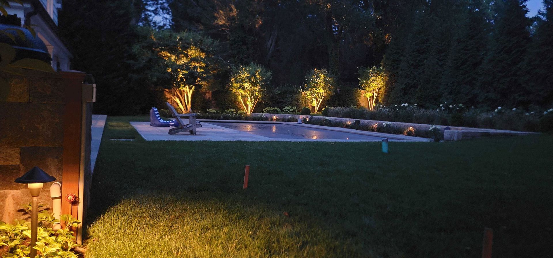 A backyard at night with a pool, trees illuminated, and lights on the grass and near a wall.
