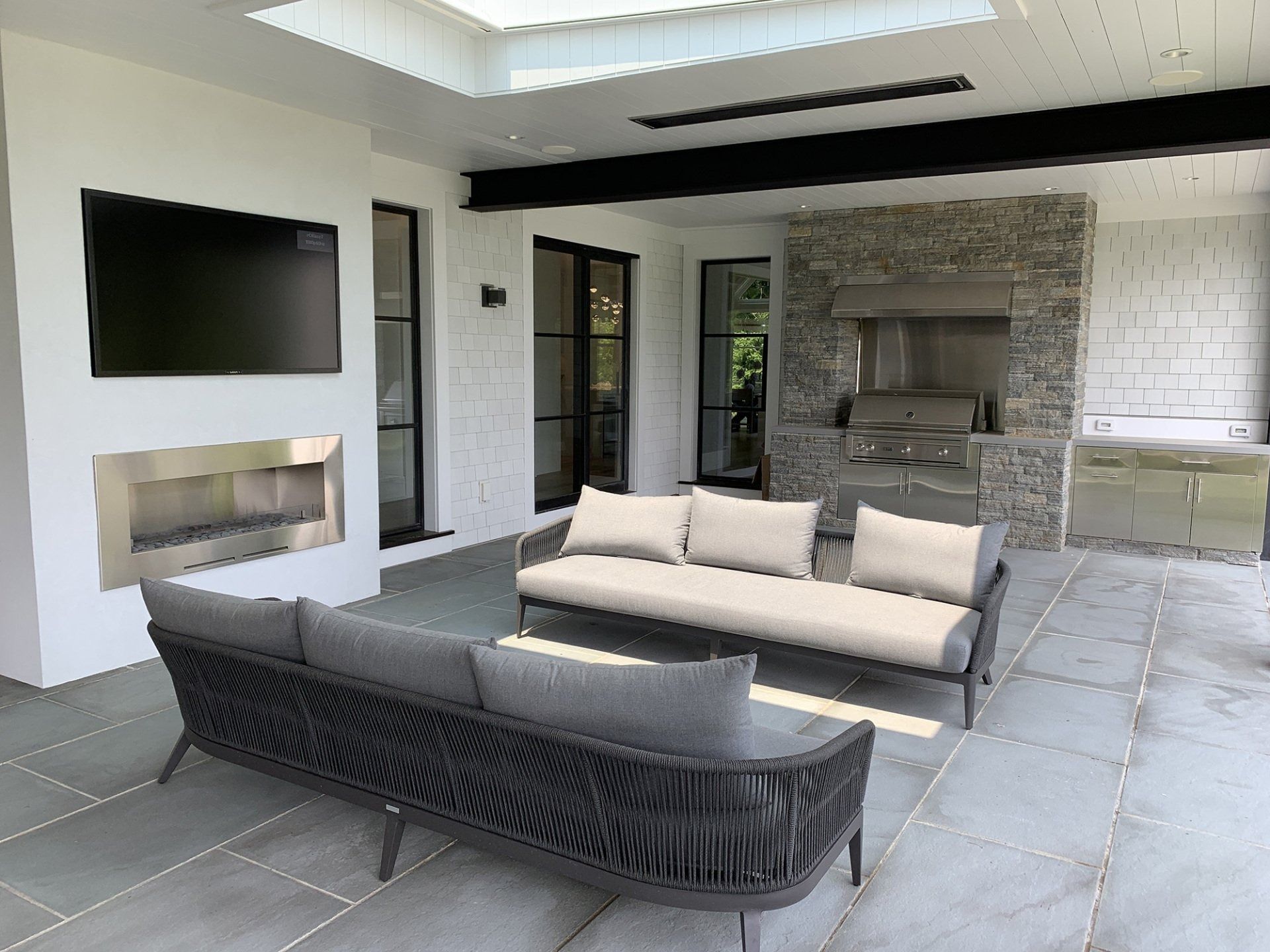Outdoor patio with gray sofas, fireplace, TV, grill, and stone wall under a skylight.