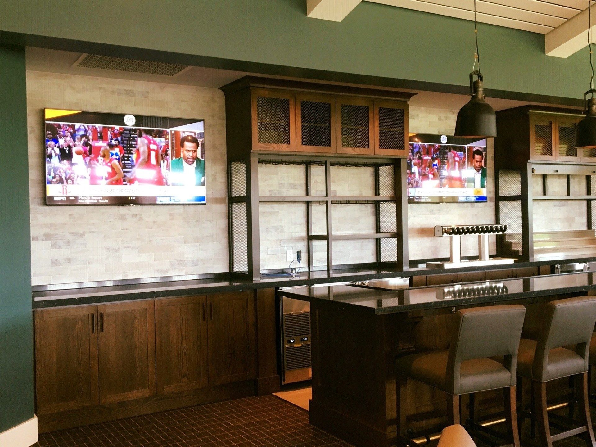 Bar with multiple TVs, wood cabinets, black bar, and stools. Two black pendant lights hang.