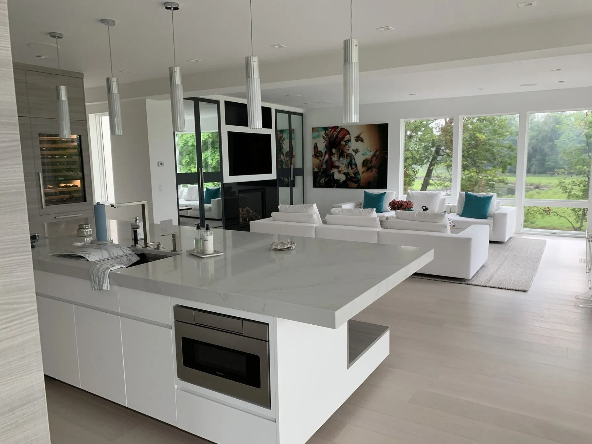 Modern white kitchen with a large island, connected to a living area with white sofas and large windows.
