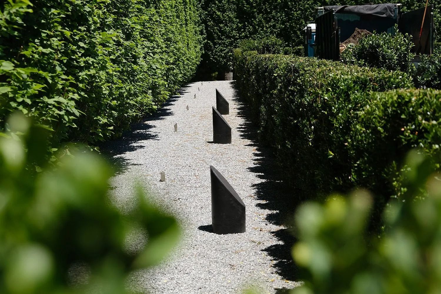 Gravel path bordered by green hedges, with three dark, angular markers. Bright sunlight.