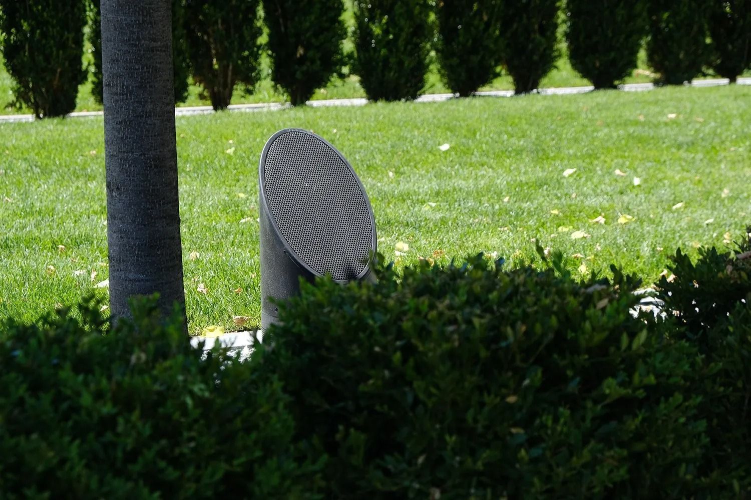 Grey lamp with woven pattern, behind green shrubs and a tree trunk, set in a grassy yard.
