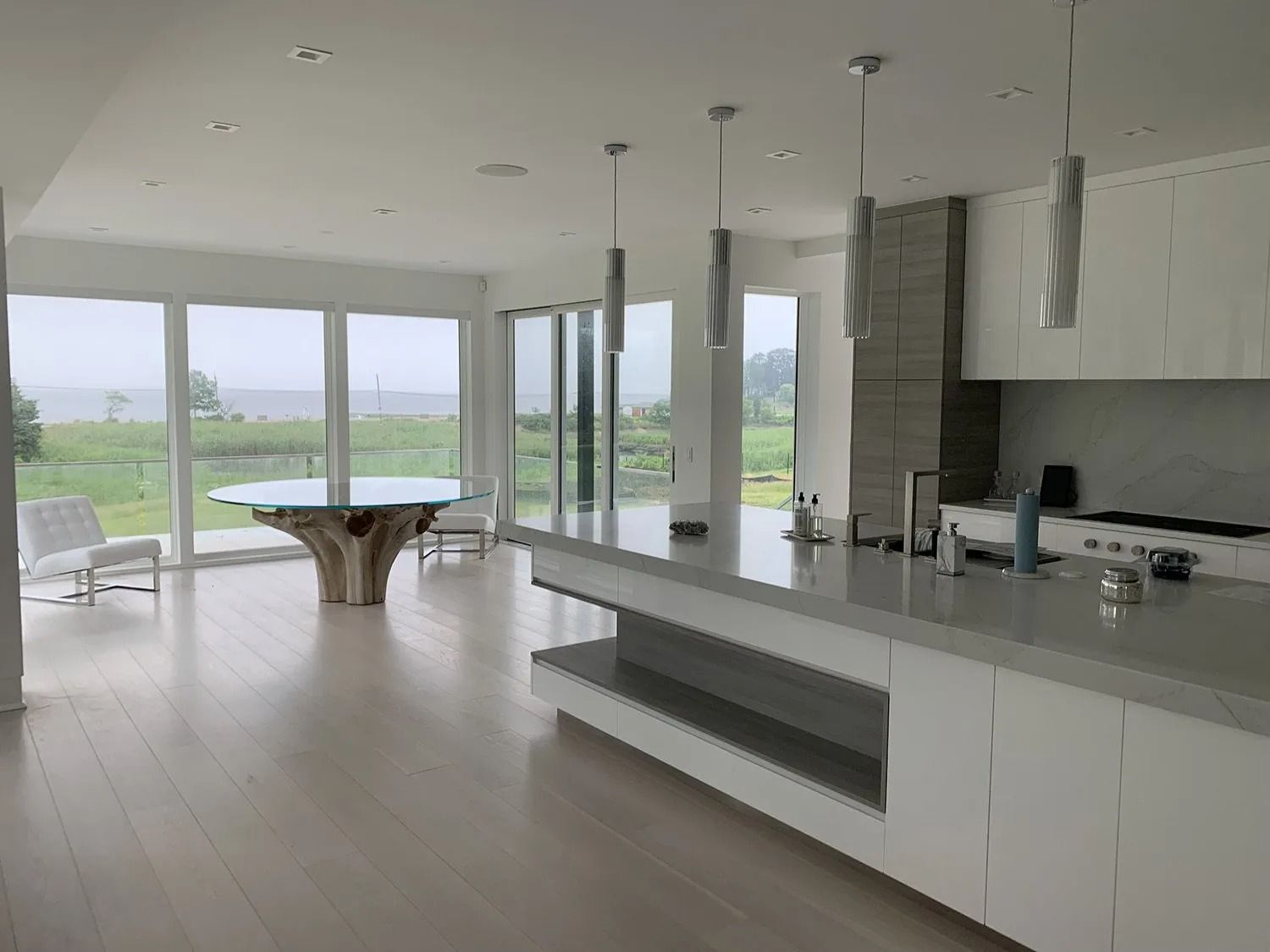 Modern, bright kitchen with large windows overlooking a field and water. White cabinets, countertops, and wood floor.